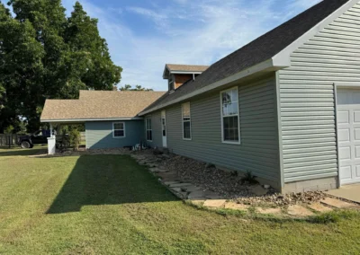 A blue house with a sloped roof, grassy yard, and a large tree nearby, featuring a garage and stone landscaping.