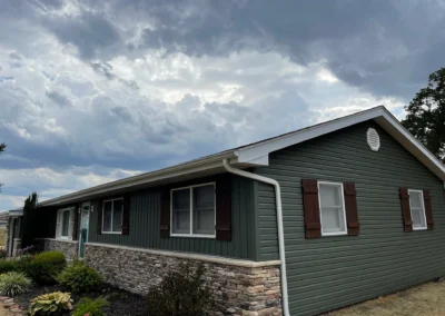 A modern home with green siding and stone accents under a dramatic sky filled with clouds, surrounded by plants and landscaping.